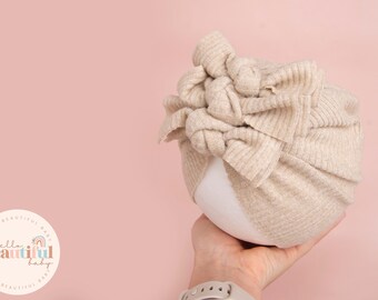 May include: A beige knitted turban headband with a large knotted bow. The headband is being held in a hand against a light pink background.