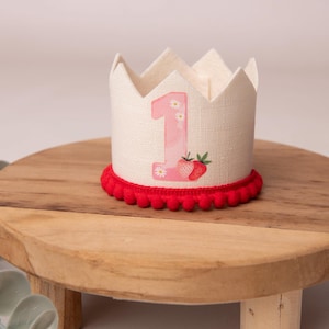 May include: A cream-colored fabric crown with a red pom-pom trim. The crown features a pink number 1 with daisy accents and a strawberry illustration. The crown is designed for a baby's first birthday.