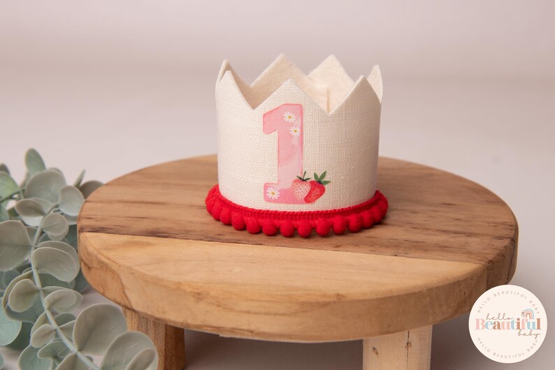 May include: A cream-colored fabric crown with a red pom-pom trim. The crown features a pink number 1 with daisy accents and a strawberry illustration. The crown is designed for a baby's first birthday.