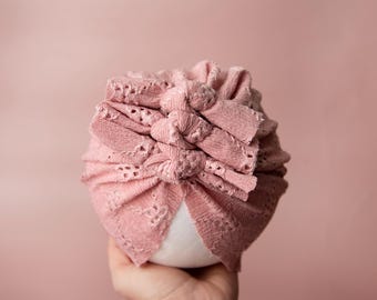 May include: A pink crocheted turban with a knotted design. The turban is made of a soft, delicate fabric and has a lace trim.