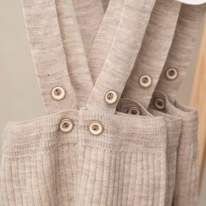 May include: Beige ribbed knit overalls with adjustable straps and wooden button closures.