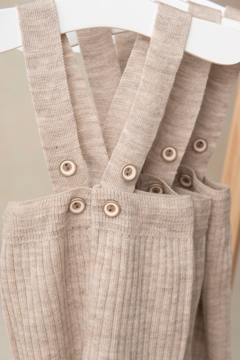 May include: Beige ribbed knit overalls with adjustable straps and wooden button closures.
