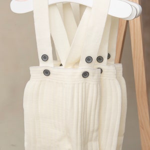 May include: Two pairs of white knitted baby overalls with suspenders and button closures. The overalls are hanging on white hangers.