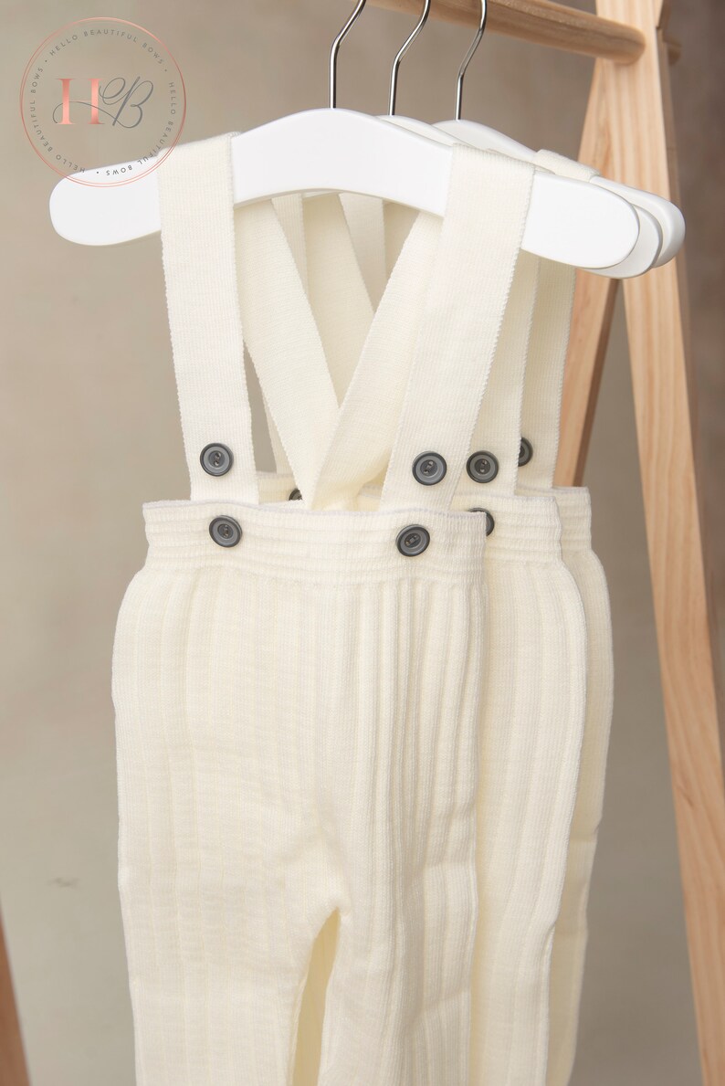 May include: Two pairs of white knitted baby overalls with suspenders and button closures. The overalls are hanging on white hangers.
