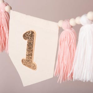Peut inclure: Guirlande de f&ecirc;te avec un num&eacute;ro 1 dor&eacute; &agrave; paillettes sur un fanion cr&egrave;me. La guirlande est orn&eacute;e de pompons roses et blancs et de perles en bois, id&eacute;ale pour une f&ecirc;te de premier anniversaire.