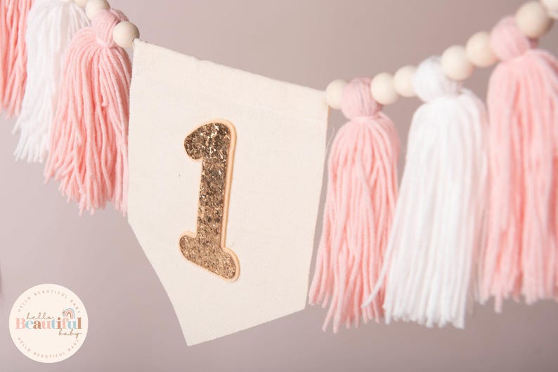 Peut inclure: Guirlande de f&ecirc;te avec un num&eacute;ro 1 dor&eacute; &agrave; paillettes sur un fanion cr&egrave;me. La guirlande est orn&eacute;e de pompons roses et blancs et de perles en bois, id&eacute;ale pour une f&ecirc;te de premier anniversaire.