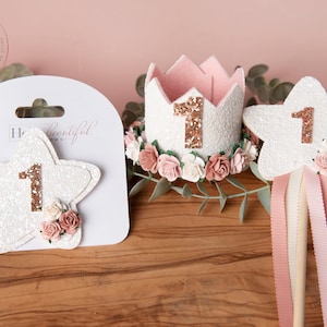 Pu&ograve; includere: Un set di tre decorazioni a forma di stella bianca con fiori rosa e un numero "1" in glitter dorato su ciascuna. Le decorazioni includono una corona, una bacchetta magica e una forcina per capelli.