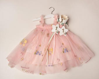 Puede incluir: Vestido de tul rosa con detalles florales, corona y varita a juego. El vestido tiene una falda en capas y está decorado con flores bordadas. Una varita en forma de estrella con el número "1" está adjunta.