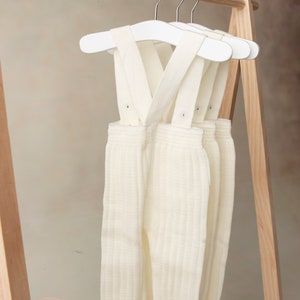 May include: Three pairs of white knitted baby overalls with suspenders hanging on white hangers. The overalls are ribbed and have a cream color.