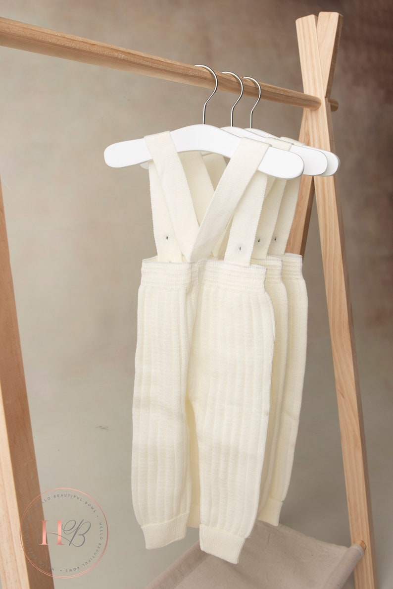 May include: Three pairs of white knitted baby overalls with suspenders hanging on white hangers. The overalls are ribbed and have a cream color.