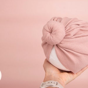 May include: A soft, ribbed knit baby turban in a dusty rose color. The turban is knotted at the top and is held in a hand against a matching pink background. The turban is designed for infants and newborns.