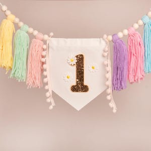 Op de afbeelding: Een kleurrijke slinger met een witte vlag met een glitterende nummer 1 en madeliefjesaccenten. De slinger is versierd met pastelkleurige garenkwasten en houten kralen, wat een vrolijke decoratie cre&euml;ert.