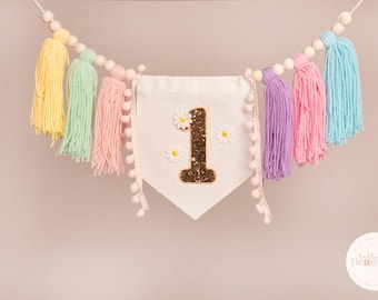 May include: A colorful banner with a white pennant featuring a glittery number 1 and daisy accents. The banner is adorned with pastel-colored yarn tassels and wooden beads, creating a cheerful and festive decoration.