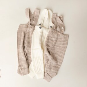 May include: Three pairs of ribbed knit baby overalls in beige and white. The overalls have adjustable straps with button closures.