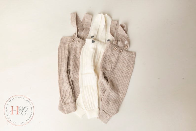 May include: Three pairs of ribbed knit baby overalls in beige and white. The overalls have adjustable straps with button closures.