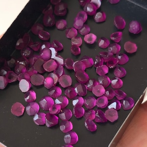 May include: A collection of small, faceted, translucent magenta-coloured gemstones scattered across a black rectangular tray. Each stone features a multi-faceted surface that reflects light, creating a vibrant, deep pink appearance against the dark background.