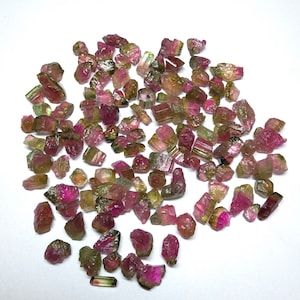 May include: A collection of pink, green and black tourmaline rough gemstones. The gemstones are various sizes and shapes, and some have a pointed or elongated shape.