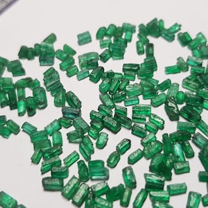 May include: A collection of small, rectangular, emerald green gemstones. The gemstones are scattered on a white surface.