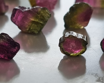 May include: A collection of small, faceted gemstones in shades of pink, green, and brown. The gemstones are arranged on a white surface.