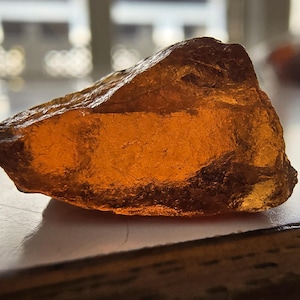 May include: A rough-cut, amber-colored gemstone with a translucent appearance. The stone has a natural, irregular shape and a warm, golden hue. The surface reflects light, highlighting its textured facets. This gemstone could be used for jewelry or as a decorative item.