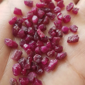 May include: A close-up image of a pile of small, red ruby gemstones, ranging in size from 3mm to 5mm. The gemstones are irregularly shaped and have a shiny, polished finish.