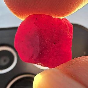 Puede incluir: Primer plano de una gema roja vibrante, sostenida entre dos dedos. La gema tiene una superficie rugosa y texturizada y un color profundo y saturado. El fondo está borroso, enfocando la atención en la gema.