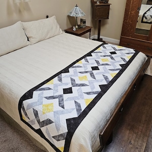 May include: A white bed with a black and gray geometric patterned bed runner with yellow accents. The bed runner has a black border.