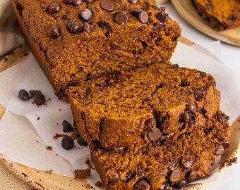 May include: A close-up of a sliced loaf of pumpkin bread with chocolate chips on a brown plate. The bread is a deep orange color and the chocolate chips are scattered throughout the bread.