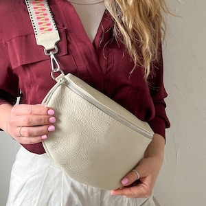 May include: A white leather fanny pack with a zipper closure and a colorful patterned strap. The strap is pink, orange, and white.