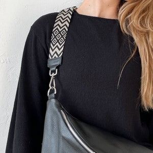 Puede incluir: Un bolso bandolera de cuero negro con una cremallera plateada y una correa con estampado en blanco y negro.