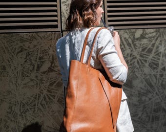 Könnte beinhalten: Eine große, braune Ledertasche mit langen Henkeln. Die Tasche wird über der Schulter getragen. Die Tasche hat eine strukturierte Oberfläche und ein schlichtes, klassisches Design. Die Person trägt einen hellfarbigen Blazer.