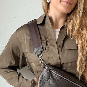 Op de afbeelding: Een bruine leren schoudertas met een bruine band en zilveren hardware. De tas wordt gedragen over een groen shirt.