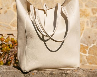 Könnte beinhalten: Cremefarbene Ledertasche mit langen Henkeln. Die Tasche hat eine rechteckige Form mit einer weiten Öffnung und einem geknoteten Detail oben. Die Tasche steht auf einer Steinoberfläche mit einer Steinmauer im Hintergrund.