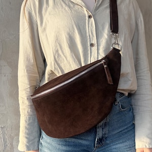 Könnte beinhalten: Eine dunkelbraune Hüfttasche aus Wildleder mit silbernem Reißverschluss und verstellbarem Riemen. Die Tasche hat eine gebogene Form und einen kleinen Leder-Reißverschlussanhänger. Der Riemen ist ebenfalls dunkelbraun.