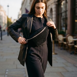 Könnte beinhalten: Eine schwarze Cord-Bauchtasche mit silbernem Reißverschluss wird über einem dunkelblauen Overall getragen. Die Person trägt eine schwarze Lederjacke. Der Hintergrund zeigt eine europäische Straßenszene.