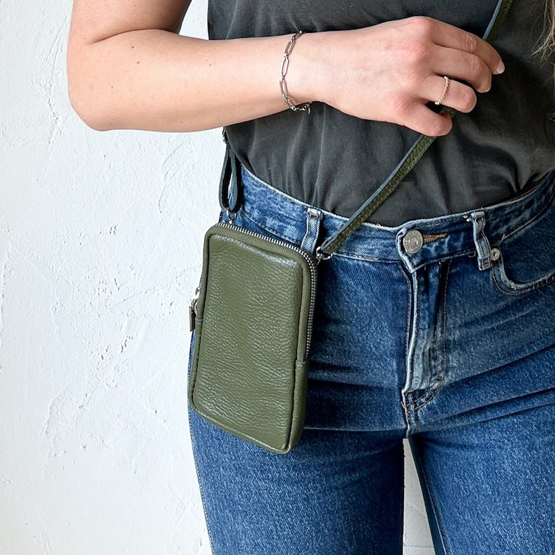 May include: A rectangular olive green leather crossbody bag with a textured surface and a silver-toned metal zipper closure. The bag features a matching thin leather strap and is worn at the hip, paired with a dark gray t-shirt and blue denim jeans.