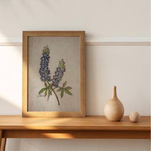 May include: A framed painting of bluebonnet flowers with green leaves on a light beige background. The painting is in a wooden frame and is hanging on a white wall.