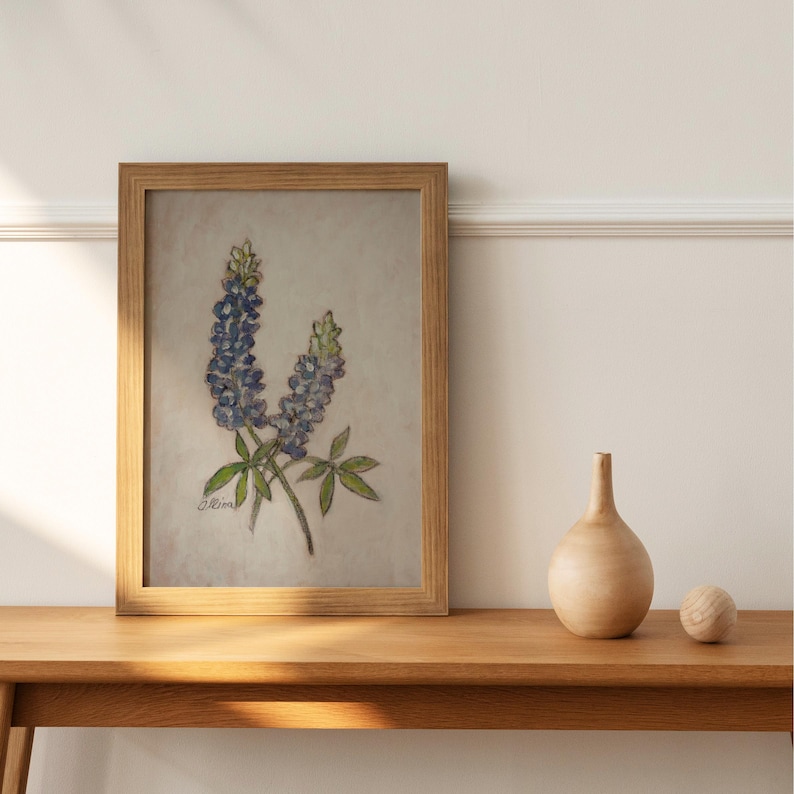 May include: A framed painting of bluebonnet flowers with green leaves on a light beige background. The painting is in a wooden frame and is hanging on a white wall.