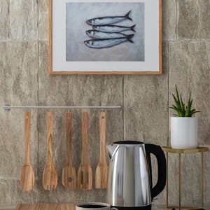 K&ouml;nnte beinhalten: Eine K&uuml;chenarbeitsplatte mit einem elektrischen Wasserkocher aus Edelstahl, einem Schneidebrett aus Holz, einem Satz Holzl&ouml;ffel, einer wei&szlig;en Tasse mit Kaffee und einem gerahmten Bild von f&uuml;nf silbernen Fischen.
