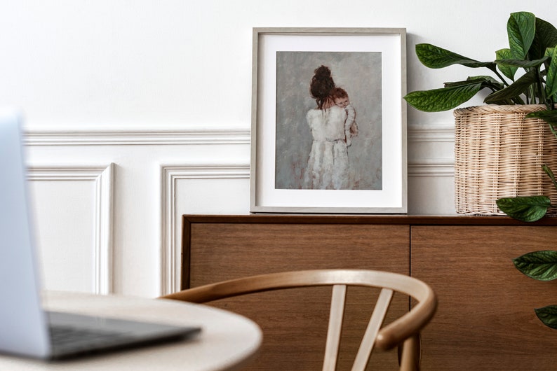 May include: A framed painting of a woman in a white dress holding a baby. The painting is on a wooden shelf with a potted plant and a laptop on a table in the foreground.