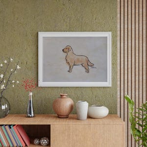 May include: A framed artwork featuring a golden retriever dog illustration. The dog is depicted in profile, with a light brown coat and a black outline. The artwork is displayed on a wall above a wooden cabinet with decorative vases and books.