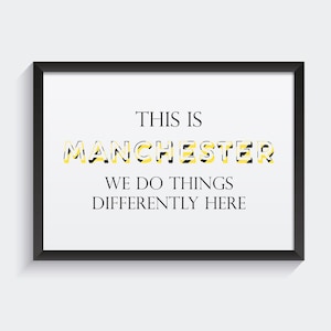 May include: A framed print with the text "THIS IS MANCHESTER WE DO THINGS DIFFERENTLY HERE." The word "MANCHESTER" is in yellow and black, with the other text in black. The frame is black.