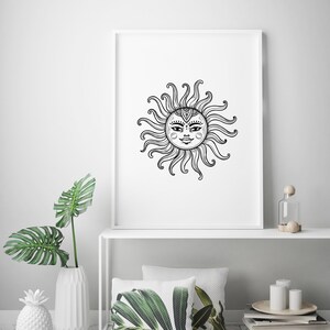 May include: A black and white illustration of a sun with a face and intricate details. The sun is framed in a white frame and is hanging on a white wall. The wall is decorated with a white vase with a green plant, a white pineapple, and a white shelf with a glass bottle, a wooden knob, and two white candles.