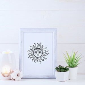 May include: A white framed print with a black and white illustration of a sun with a face. The print is on a white wooden surface with a white ceramic oil diffuser and a cotton boll.