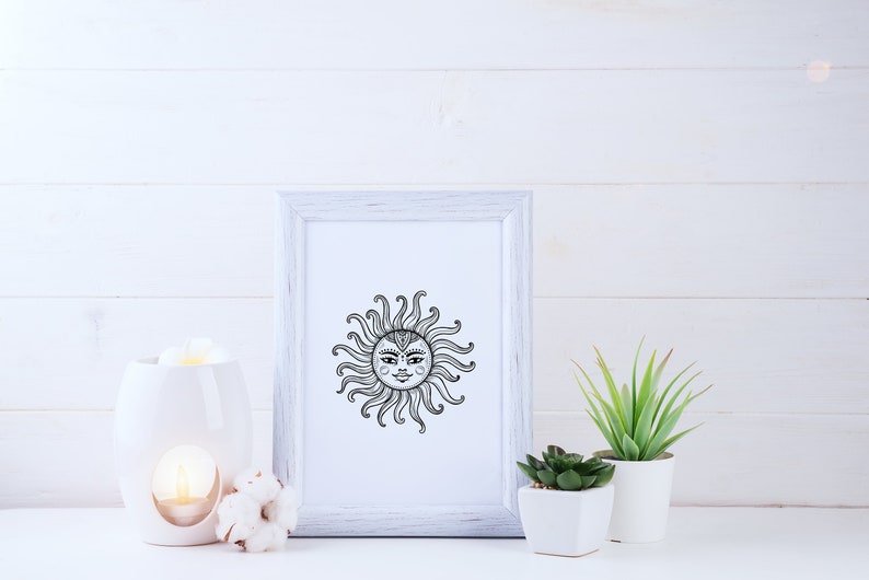May include: A white framed print with a black and white illustration of a sun with a face. The print is on a white wooden surface with a white ceramic oil diffuser and a cotton boll.