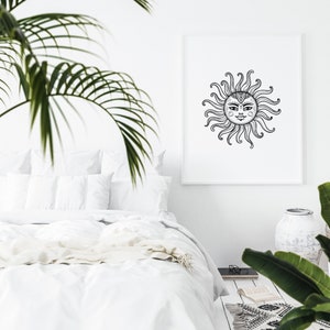 May include: A black and white illustration of a sun with a face, surrounded by rays, printed on a white poster in a white frame. The poster is hanging on a white wall above a white bed with white bedding.