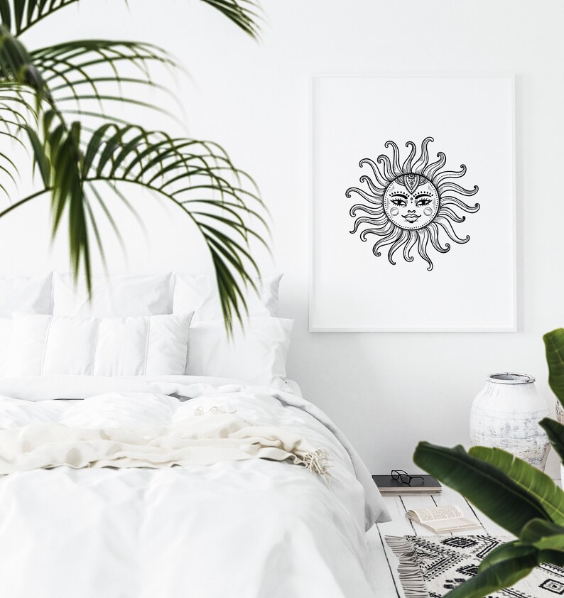 May include: A black and white illustration of a sun with a face, surrounded by rays, printed on a white poster in a white frame. The poster is hanging on a white wall above a white bed with white bedding.