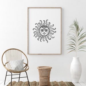 May include: A black and white line drawing of a sun with a face, surrounded by rays, in a wooden frame on a white wall. A wicker chair with a white pillow sits in front of the frame, and a wicker side table is to the right of the chair. A potted palm plant is in the background.