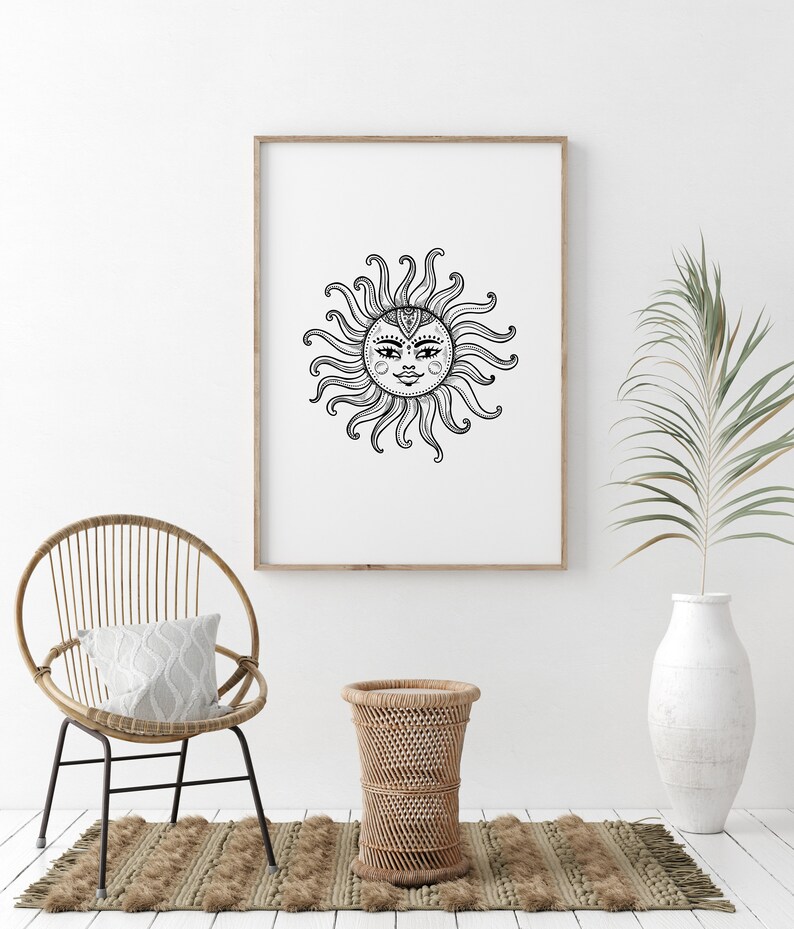 May include: A black and white line drawing of a sun with a face, surrounded by rays, in a wooden frame on a white wall. A wicker chair with a white pillow sits in front of the frame, and a wicker side table is to the right of the chair. A potted palm plant is in the background.