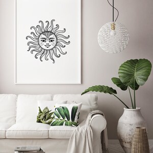 May include: A black and white illustration of a sun with a face, framed and hanging on a white wall above a white couch with green leaf patterned throw pillows.